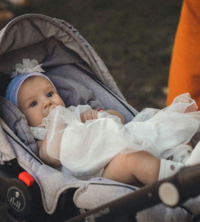 Małe dziecko leżące w czystym, szarym wózku dziecięcym, ubrane w białą sukienkę i ozdobną opaskę.